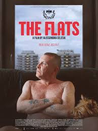 Text on poster: “The Flats.” The image shows a man sitting indoors near a window.